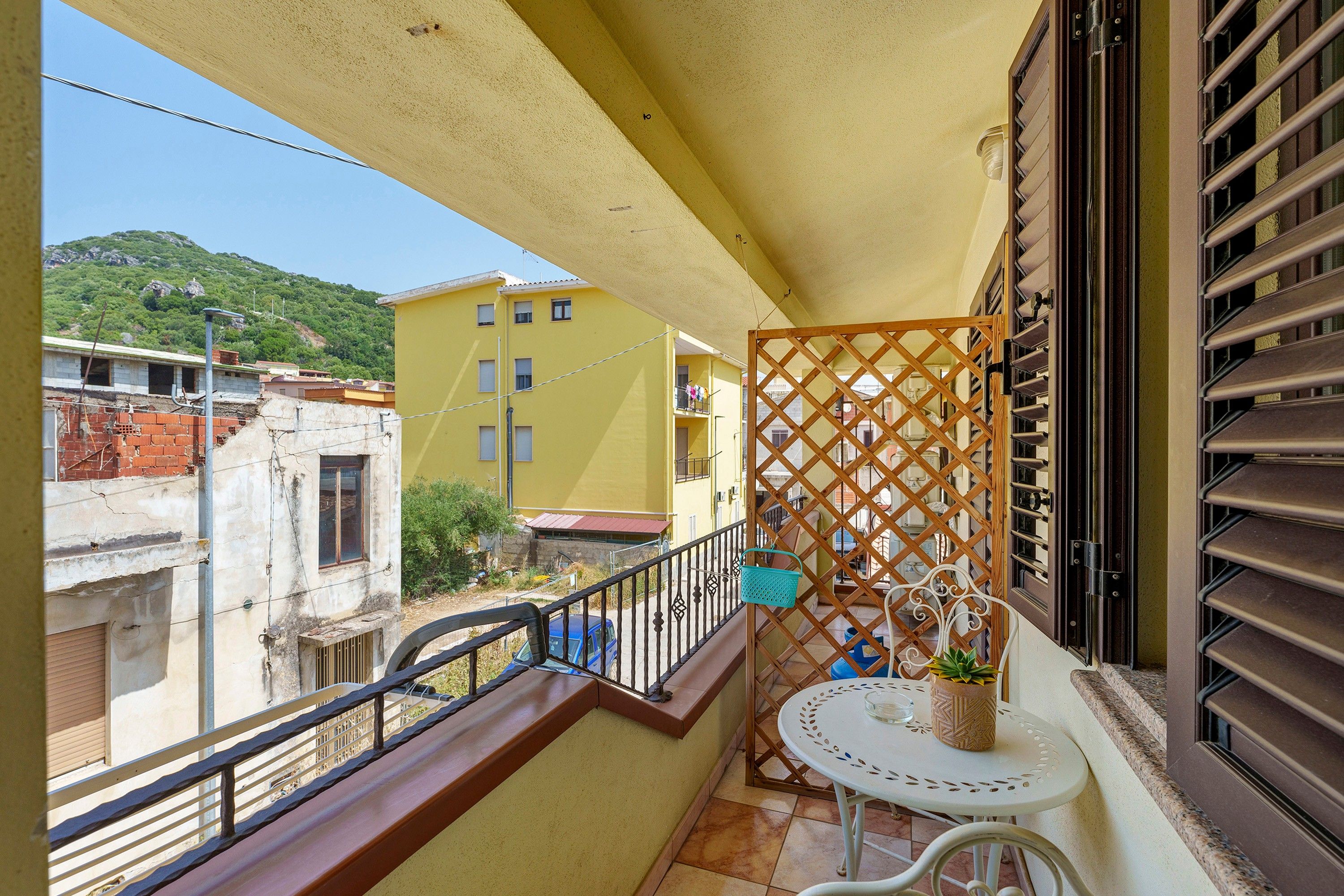 Un balcon agrémenté d’une séparation en treillis de bois, une petite table ronde avec un pot de fleurs, et une vue sur un immeuble jaune et une colline.
