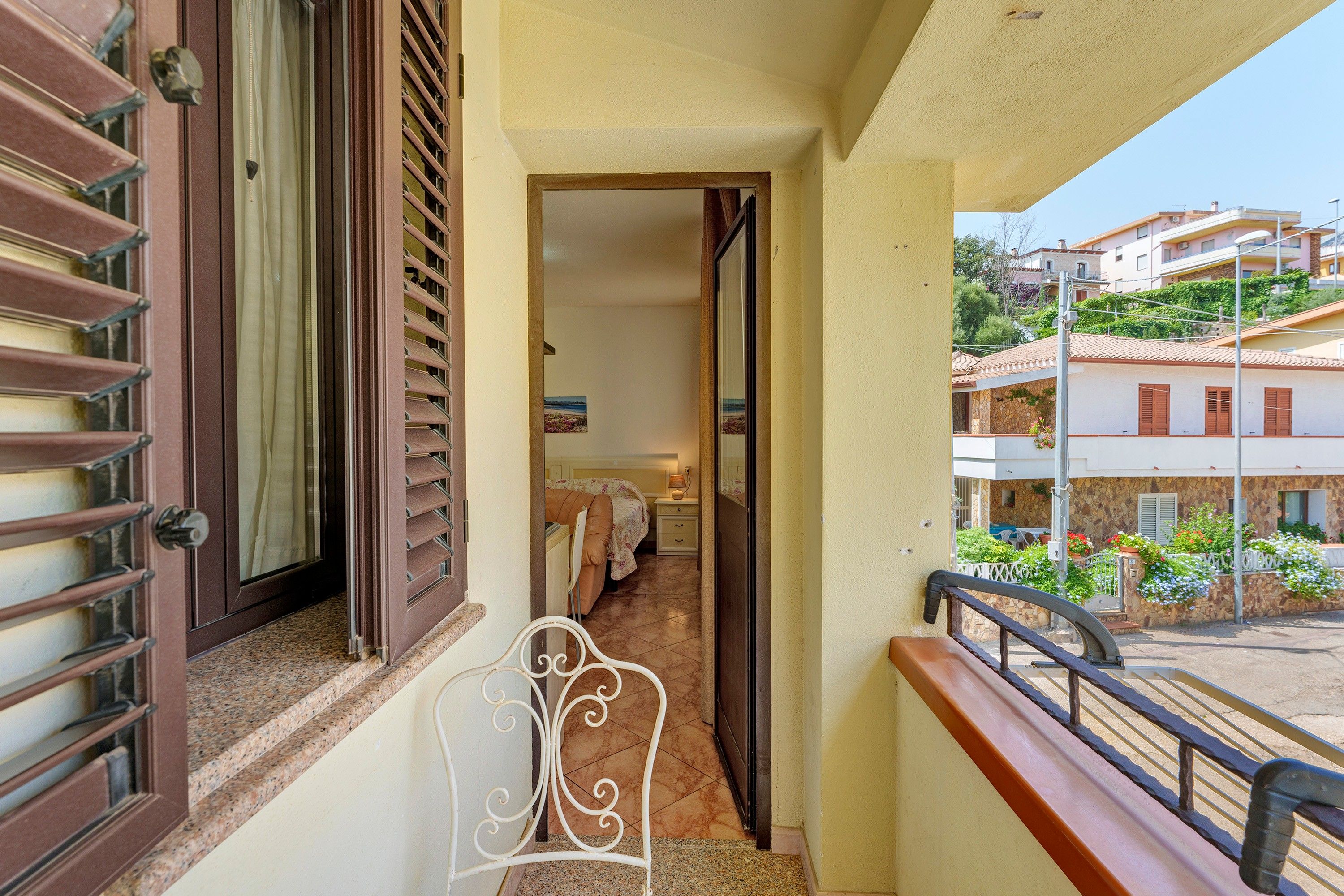 Un balcón con vistas a una zona residencial, una puerta que da a un dormitorio y una silla metálica blanca.