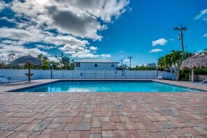 Une piscine rectangulaire bordée d’une terrasse en briques, entourée d’une clôture blanche, sous un ciel partiellement nuageux.