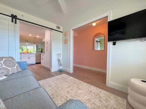 A living room with a sofa, a television mounted on the wall, and a kitchen in the background. - Sea Grape Bay #205 Bayfront Island Condo (Fort Myers Beach)