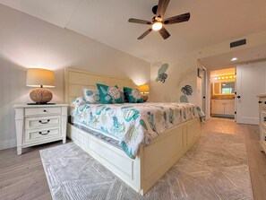 Ein Schlafzimmer mit einem Bett, Nachttischen, einem Deckenventilator und einem durchgehenden Türrahmen sichtbaren Badezimmer.