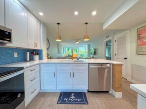 A modern kitchen with white cabinets, a built-in oven, a microwave, and a dishwasher. There is a blue hexagonal backsplash, a welcome mat, and pendant lights. - Sea Grape Bay #205 Bayfront Island Condo (Fort Myers Beach)