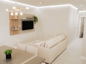 A modern living room with a sofa, a flat-screen TV, a dining table, and a plant. - 30m from the beach • 2 Suites • Grill • Garage (Porto Belo)