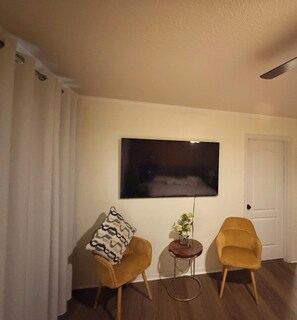 Ein Zimmer mit einem an der Wand befestigten Flachbildfernseher, zwei Sesseln, einem kleinen Beistelltisch mit einer Pflanze und einer Tür.