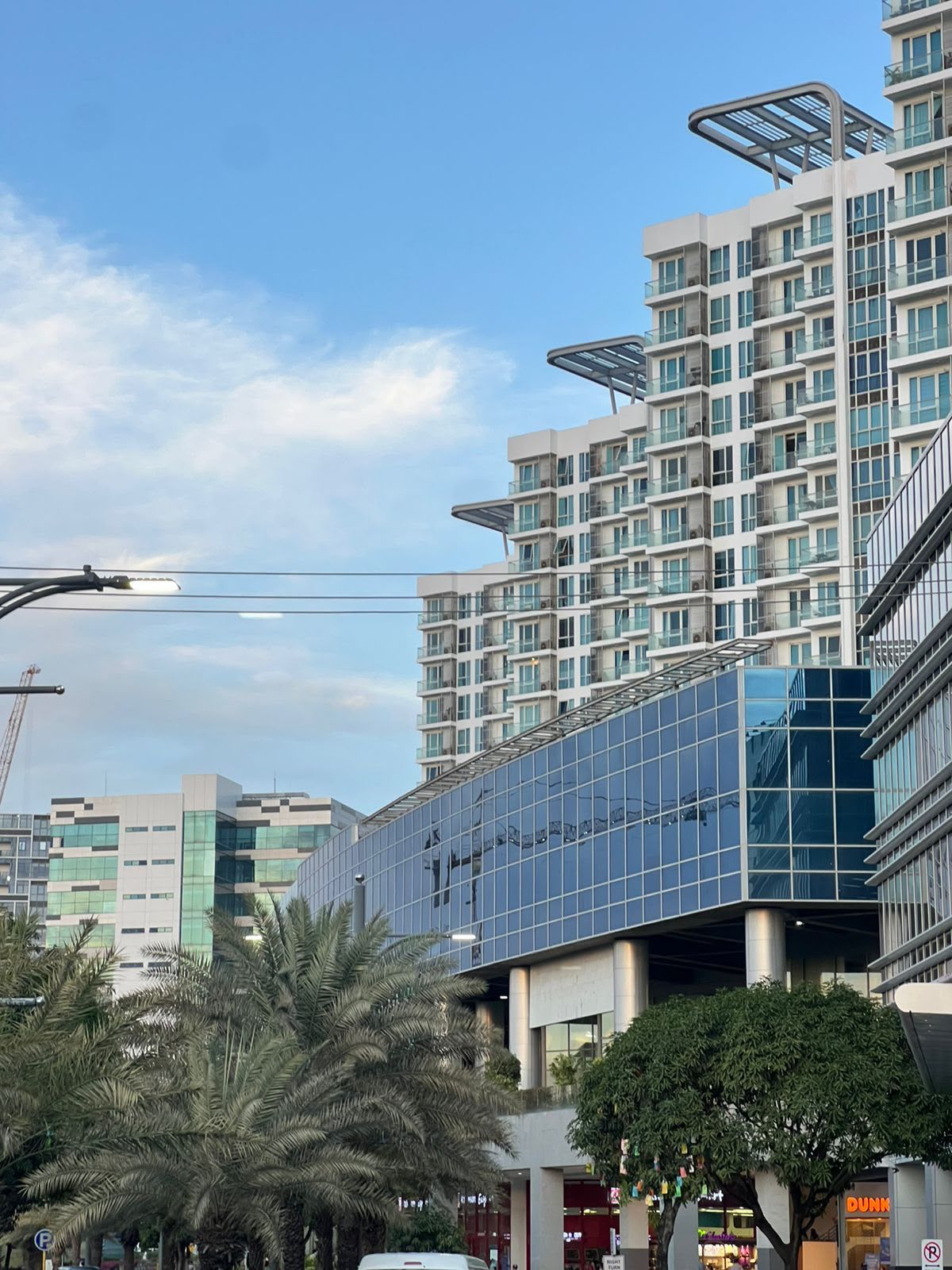A modern multi-story building with a glass facade, surrounded by palm trees and other commercial buildings.
