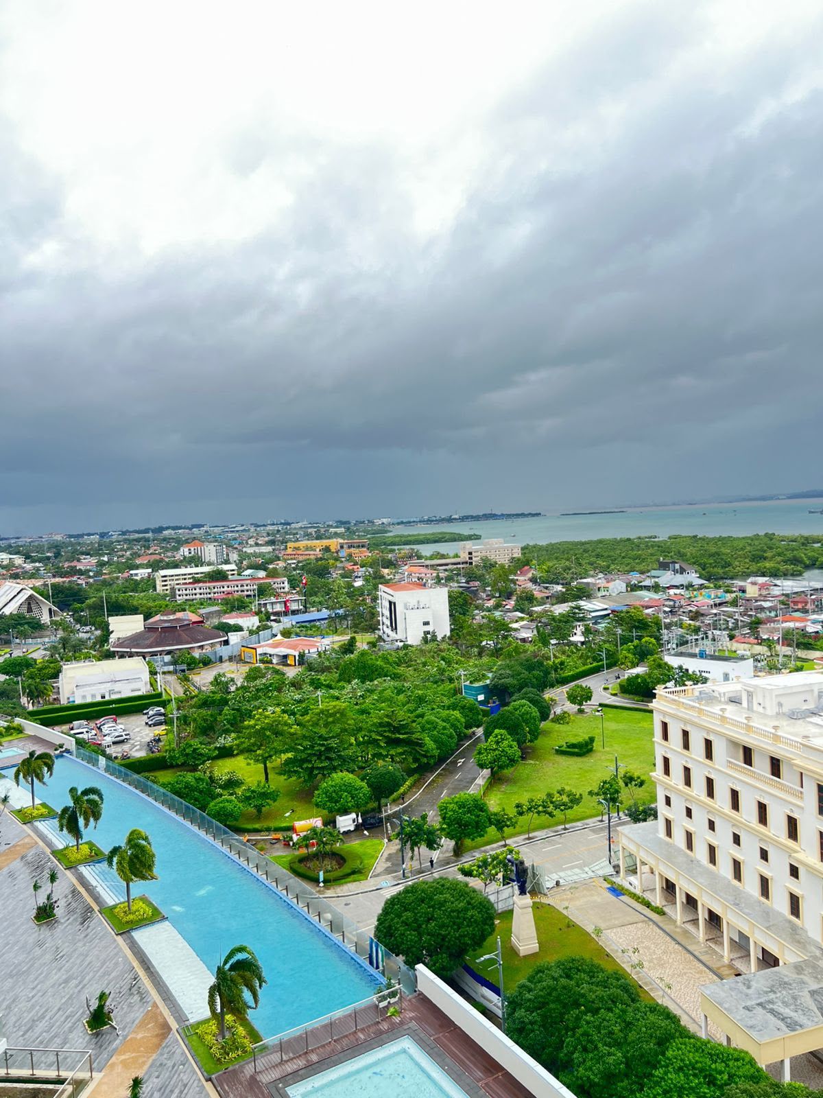 A large hotel with a swimming pool, surrounded by greenery and a view of the cityscape.