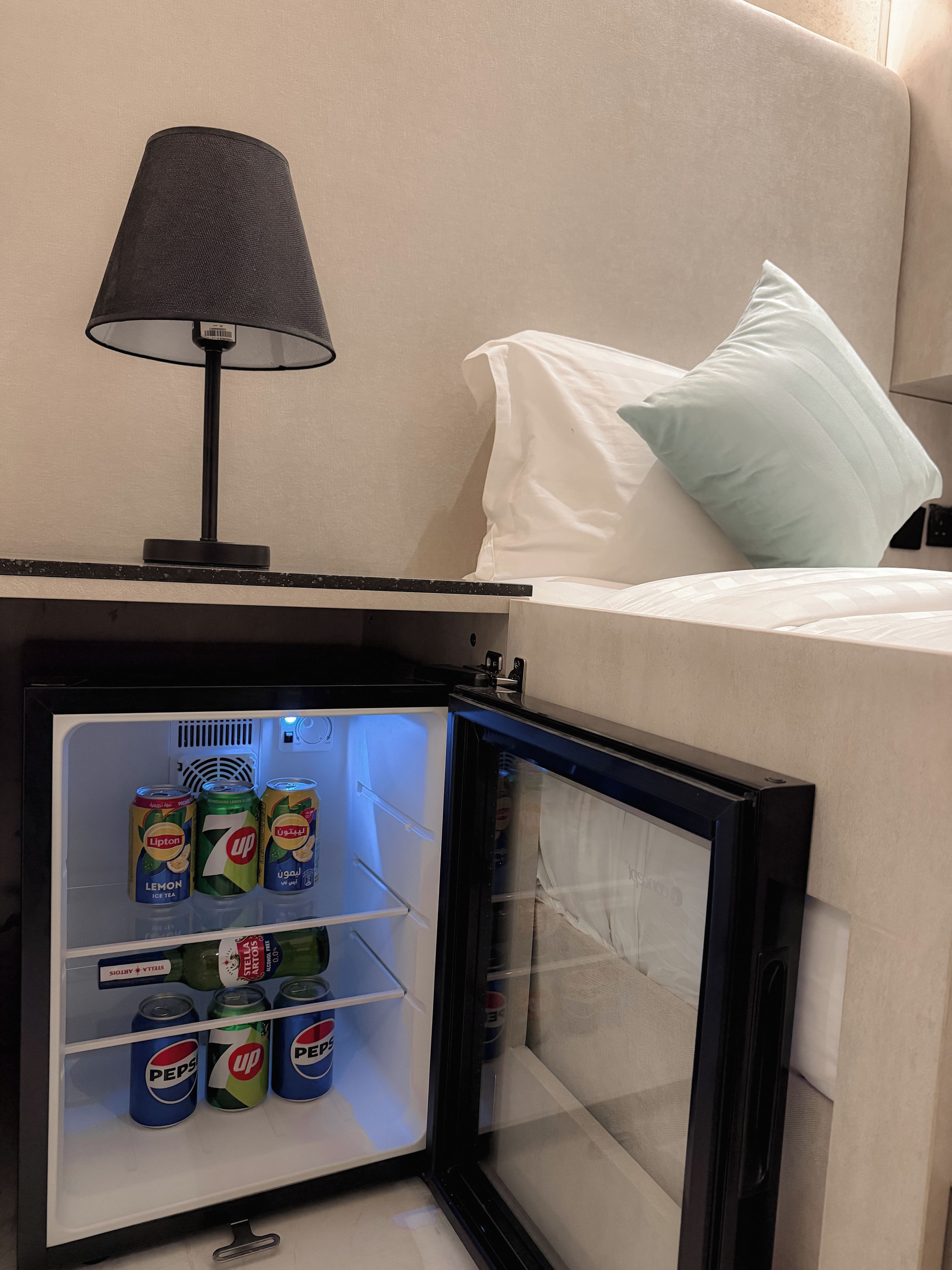 Ein Minikühlschrank mit Getränken, eine Lampe und ein Bett mit Kissen.
