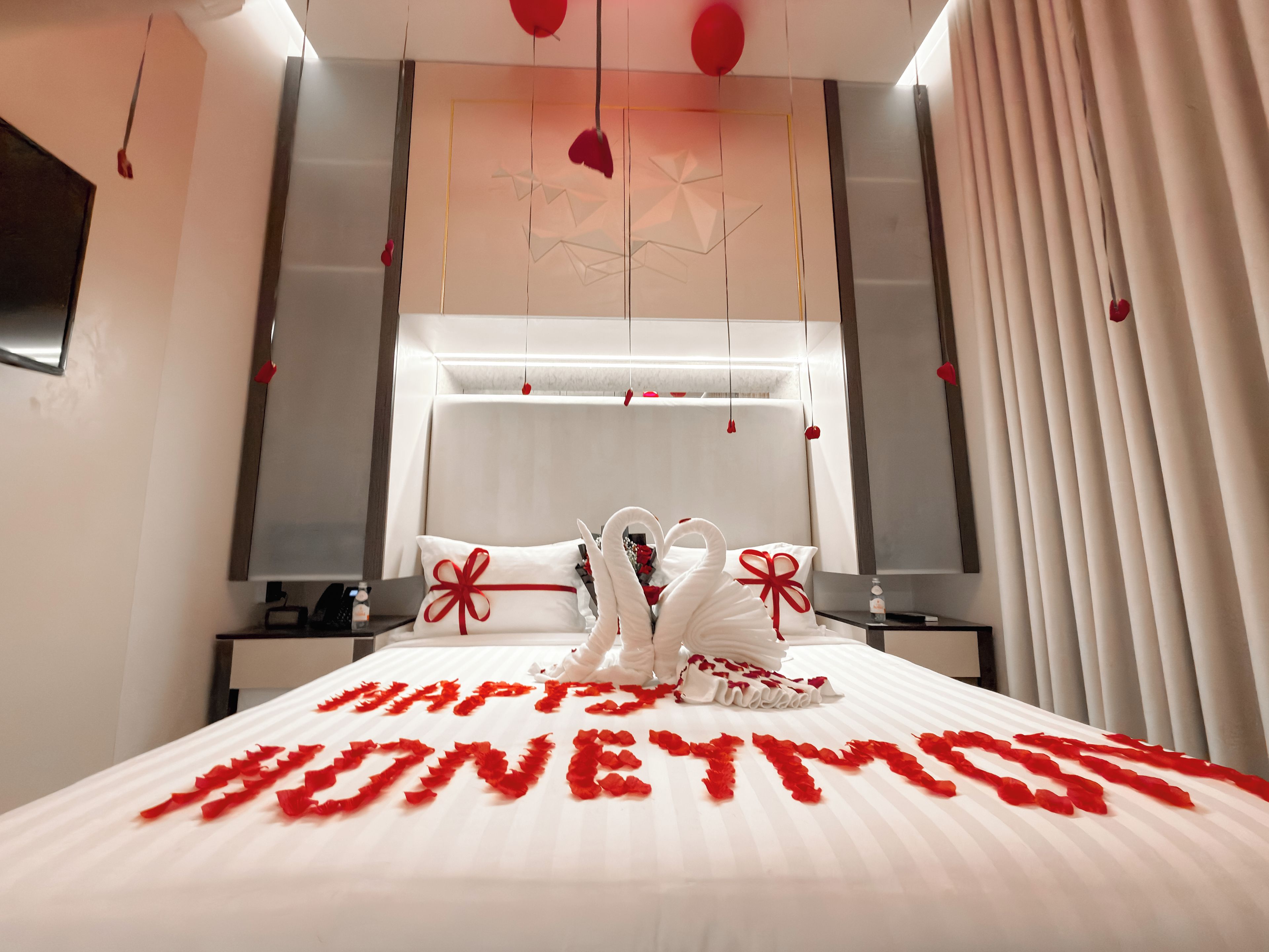 A hotel room with a bed adorned with rose petals spelling 'HONEYMOON' and two swan-shaped pillows.