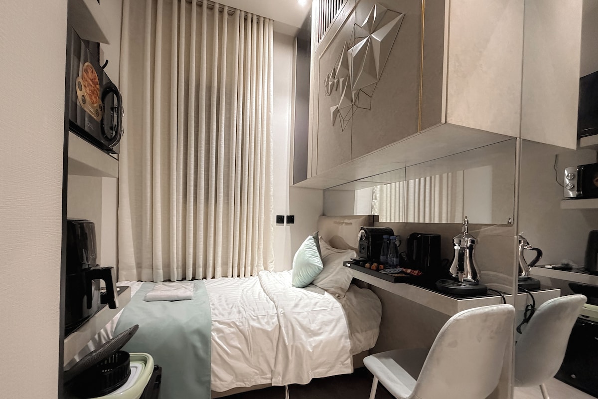 A compact hotel room with a bed, a desk, a chair, a coffee machine, and a microwave.