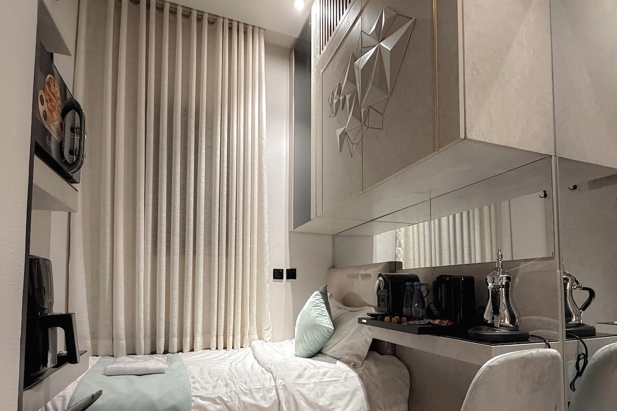 A modern hotel room with a bed, a desk, and a chair. There is a window with curtains, a wall-mounted clock, and a decorative wall panel.