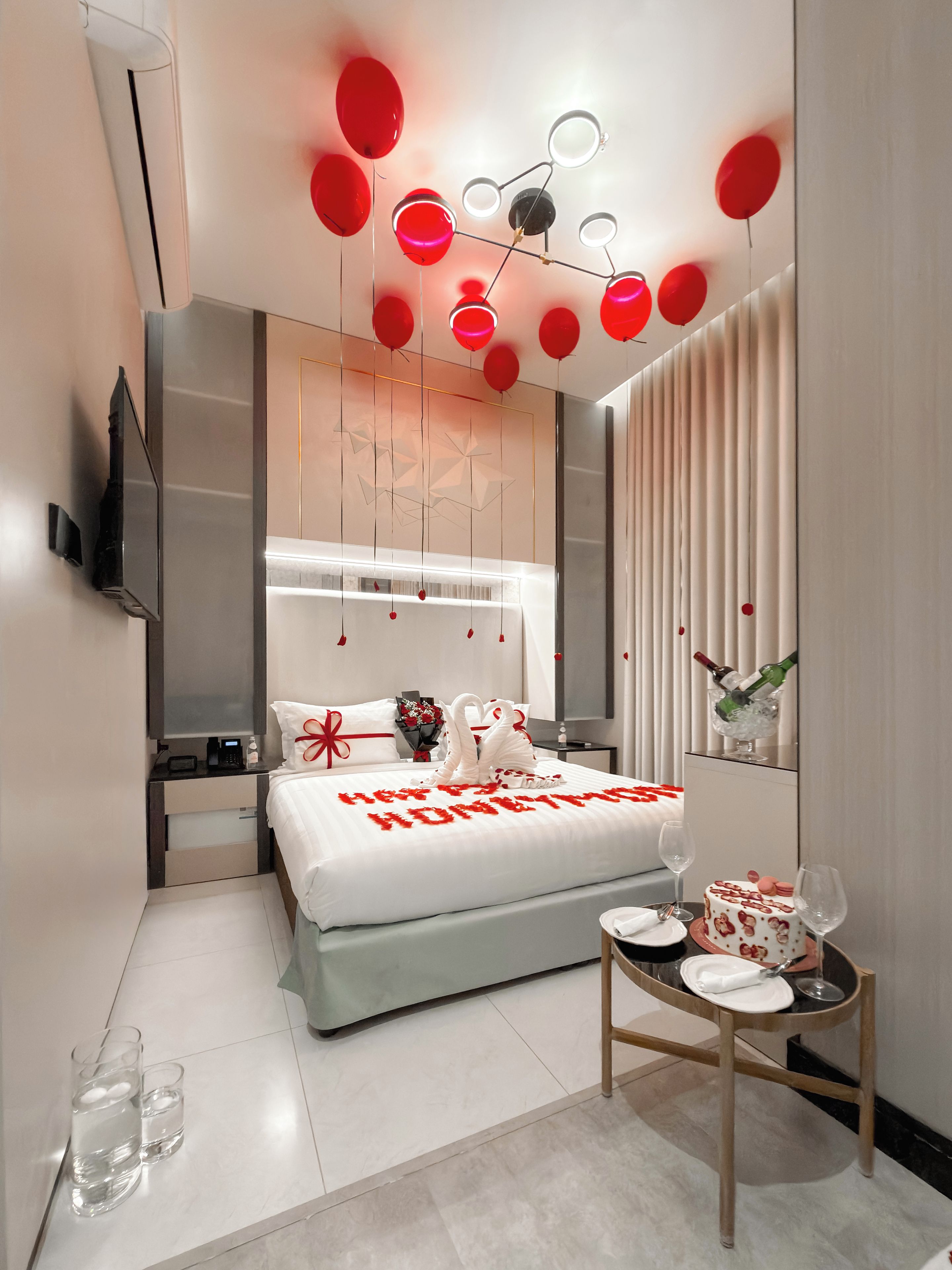 A modern hotel room with a neatly made bed, a bedside table with a glass of water, a cake, and a bouquet of flowers.