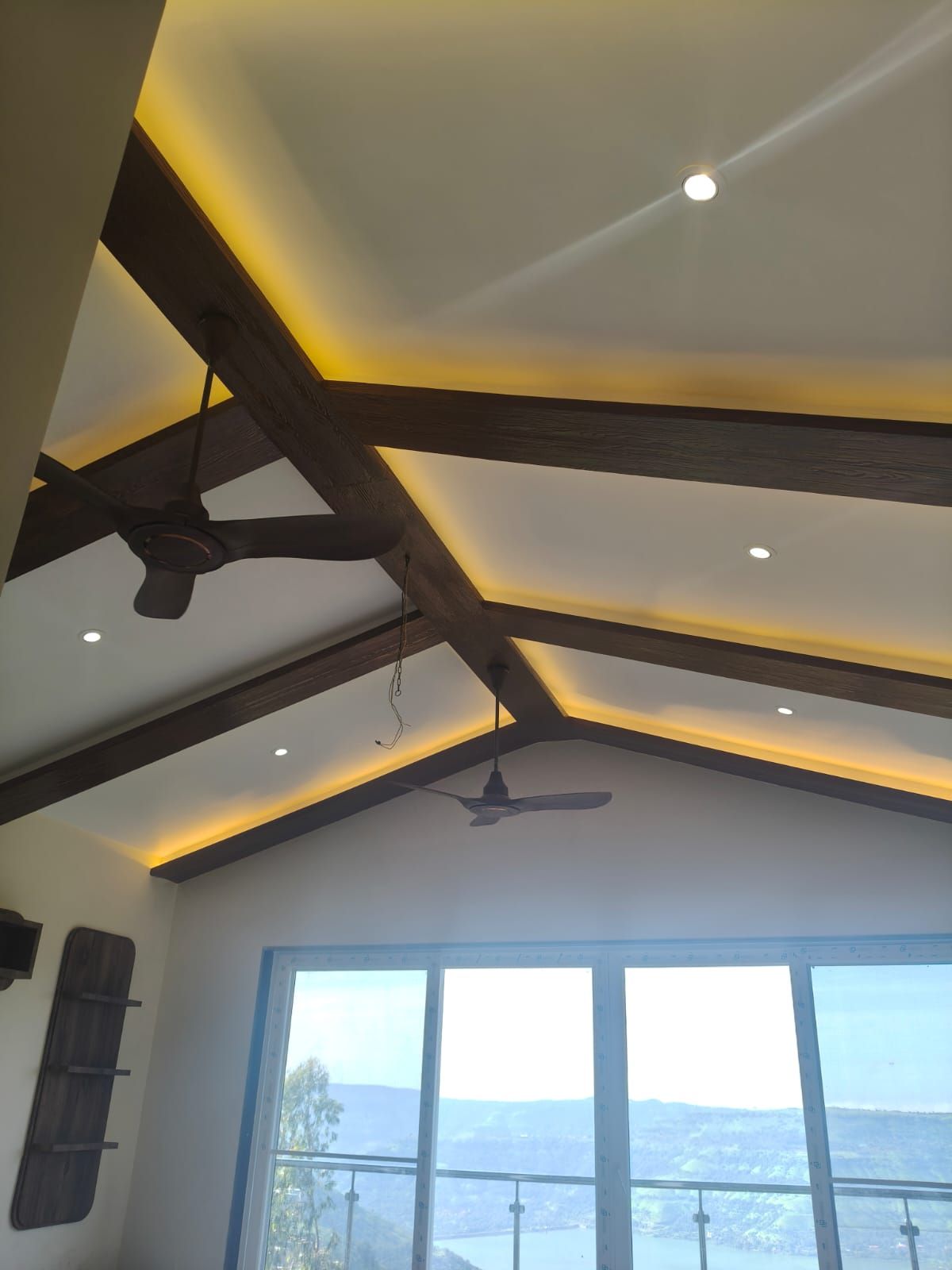 Une pièce dotée d’un plafond en pente, de poutres apparentes, d’un ventilateur de plafond, d’un éclairage encastré et d’une grande fenêtre donnant sur un paysage.