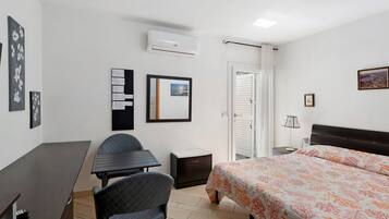 A hotel room with a bed, a desk, a chair, a small table, a nightstand, an air conditioner, a door, and a window.