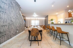 A modern dining area with a long table, multiple chairs, and a kitchen in the background. - 5BR Paradise Palms Toy Story & Minions Rooms (Kissimmee)