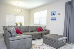 Ein Wohnzimmer mit einem grauen Ecksofa, einem grauen Hocker, einem gemusterten Teppich und einer Kronleuchter.