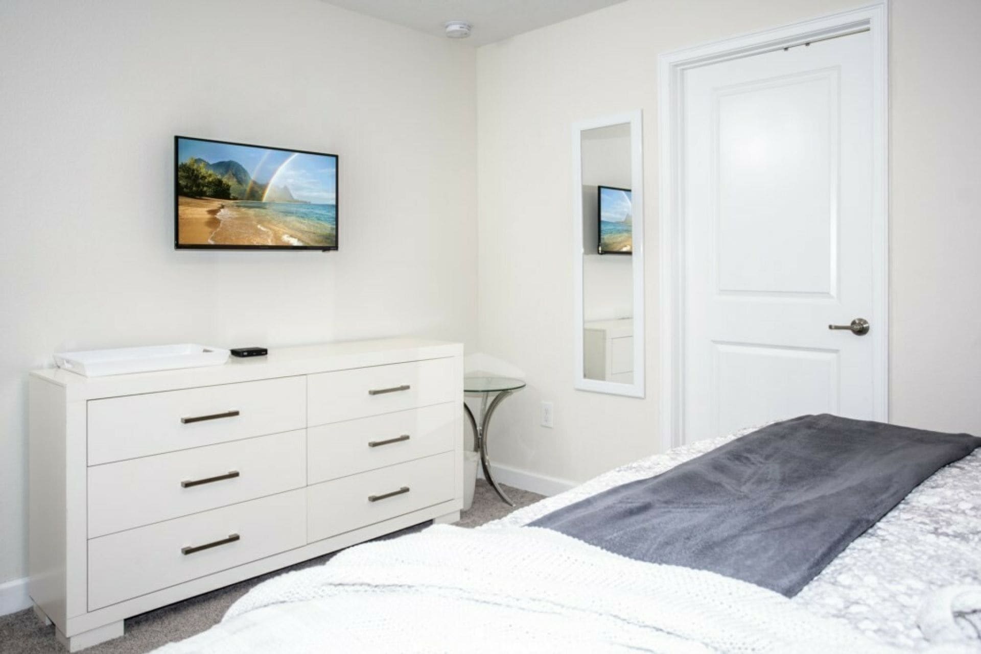 A bedroom with a bed, a dresser, a mirror, and a wall-mounted TV.