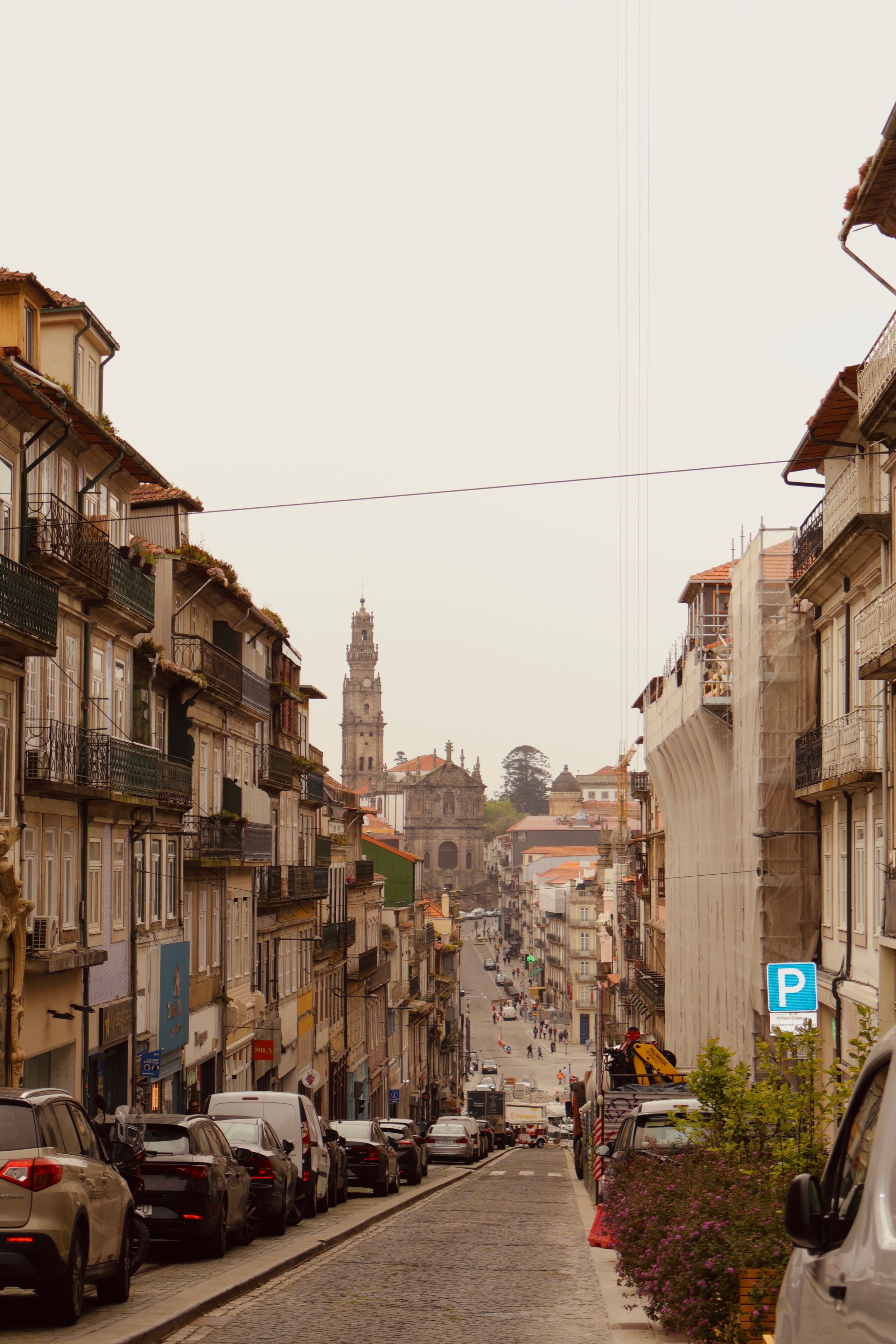 Vista da rua com edifícios, carros estacionados e uma torre de igreja imponente ao fundo.