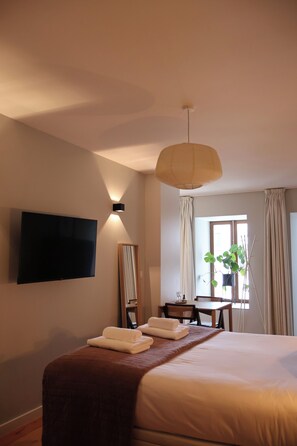 Ein Hotelzimmer mit Bett, Flachbildfernseher an der Wand, Schreibtisch mit Stuhl, Fenster mit Vorhängen und einer hängenden Pflanze.