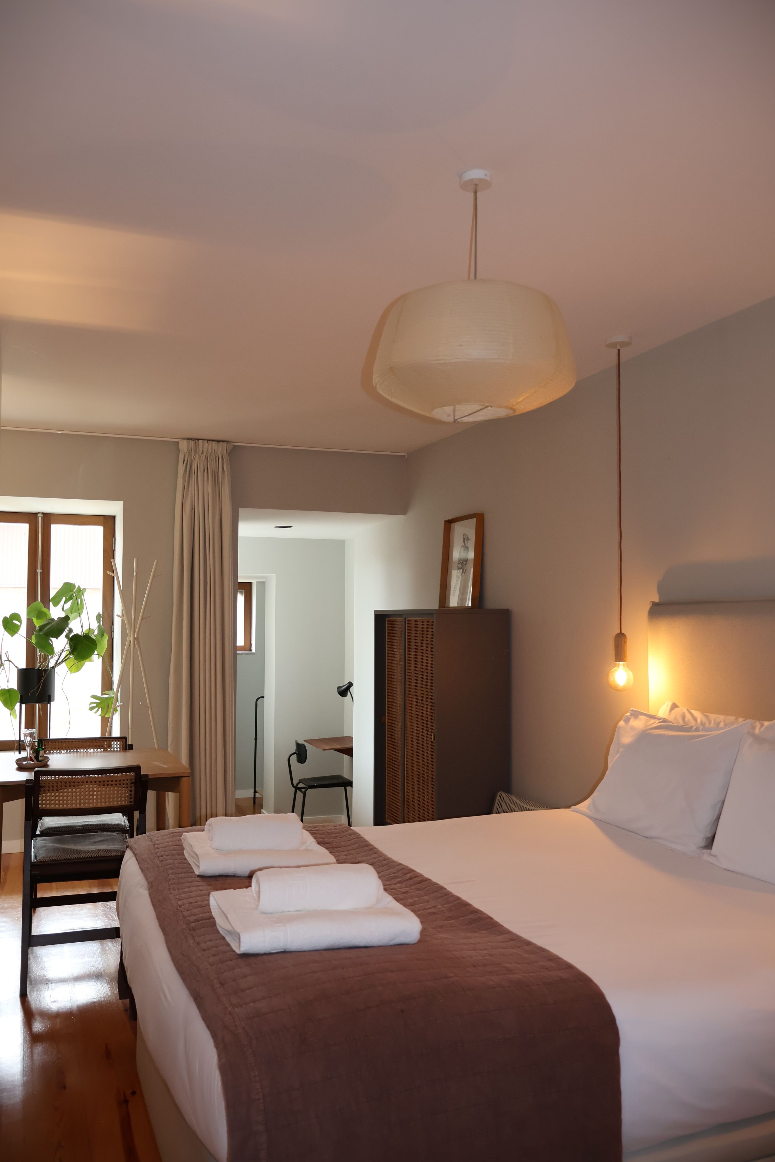 Ein Hotelzimmer mit einem großen Bett, einem Schreibtisch, einem Sessel, einer Pflanze und Blick auf den Flur.
