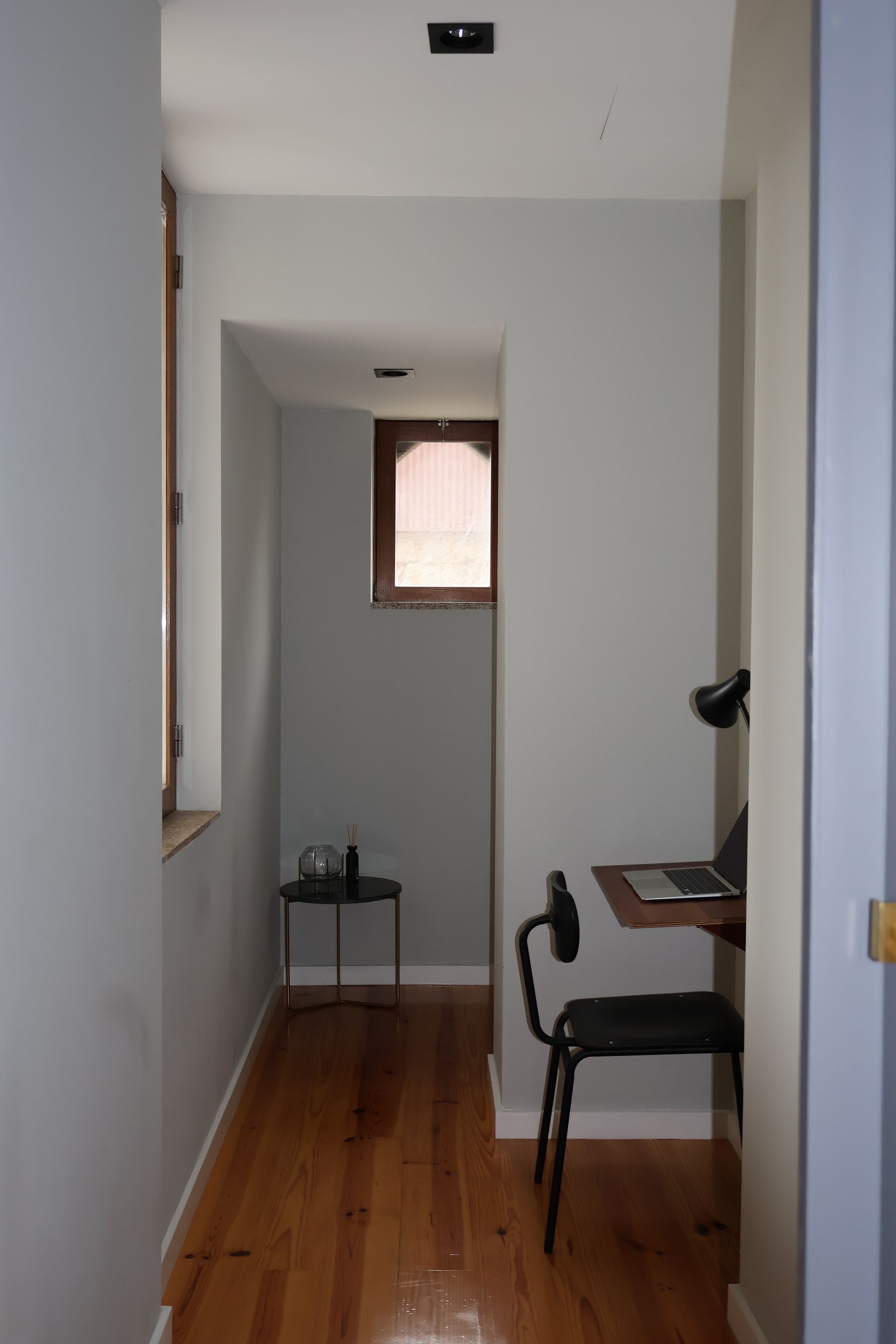 Quarto com piso de madeira, uma escrivaninha com cadeira, uma mesinha de centro com abajur e uma janela com cortina.