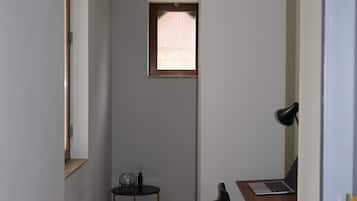 Quarto com piso de madeira, uma escrivaninha com cadeira, uma mesinha de centro com abajur e uma janela com cortina.