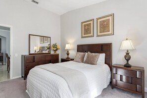 A bedroom with a large bed, a nightstand with a lamp, a dresser, a mirror, and two framed floral paintings on the wall. - 4BR Windsor Palms | Pool, spa & Mickey room (Kissimmee)