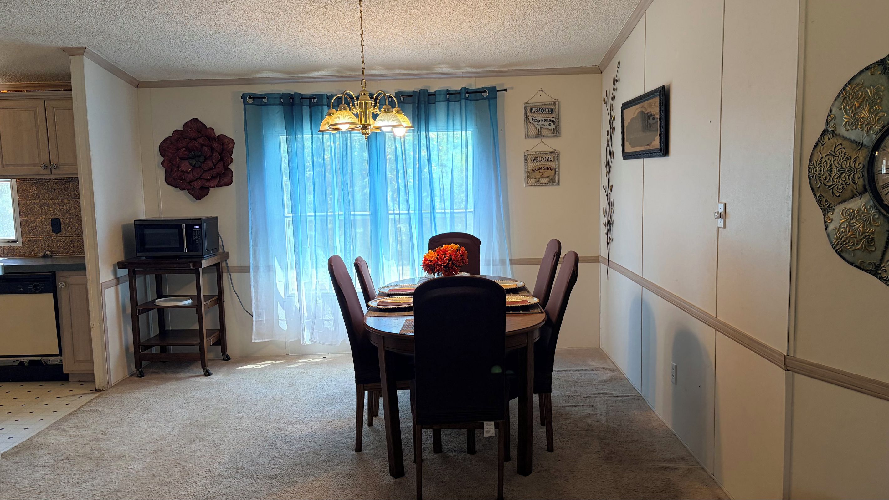 Comedor con mesa dispuesta para cuatro personas, una araña y vistas a través de una ventana con cortinas traslúcidas.
