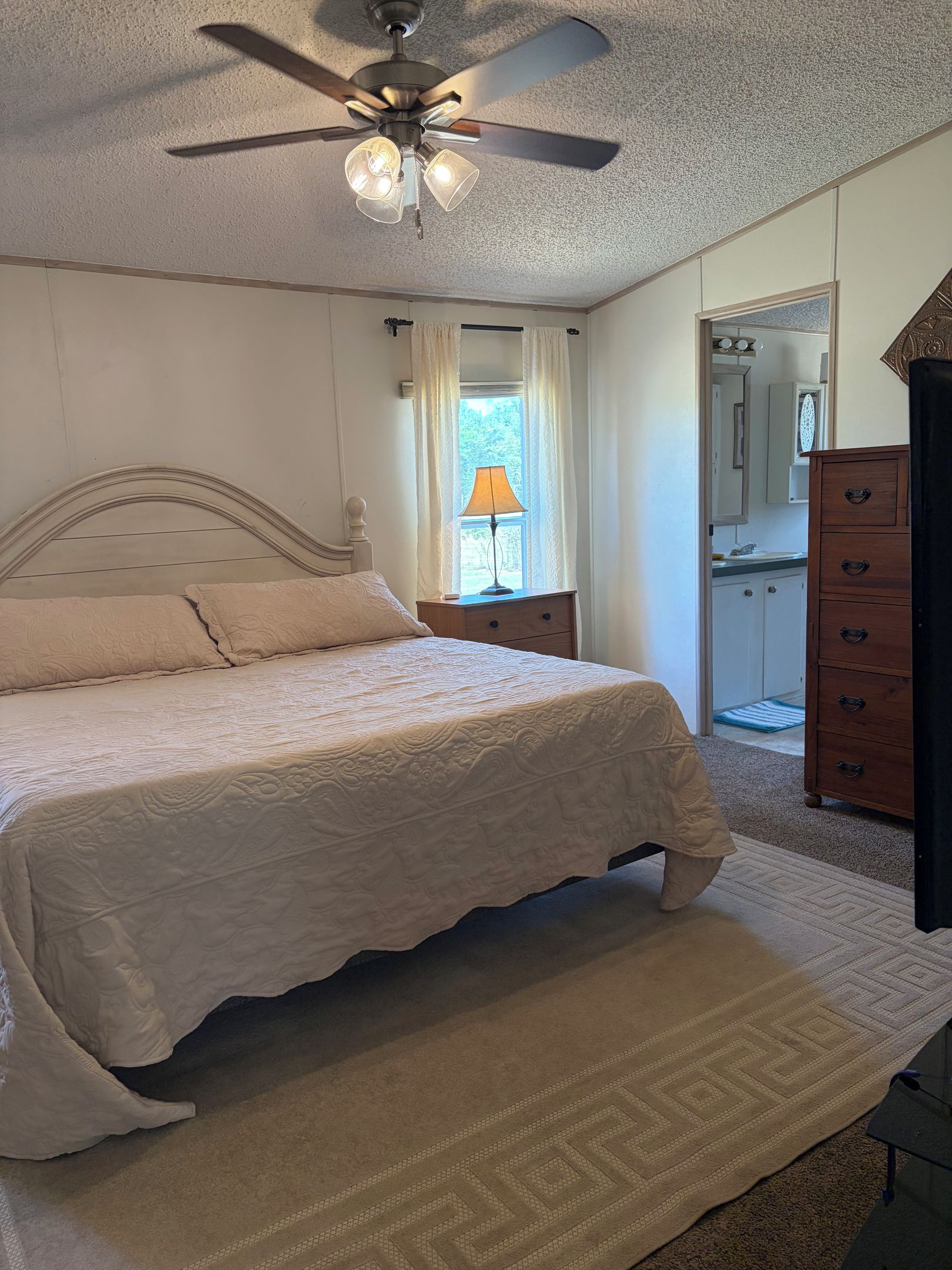 Un dormitorio con cama, ventilador de techo, mesita de noche, cómoda y ventana con cortinas.