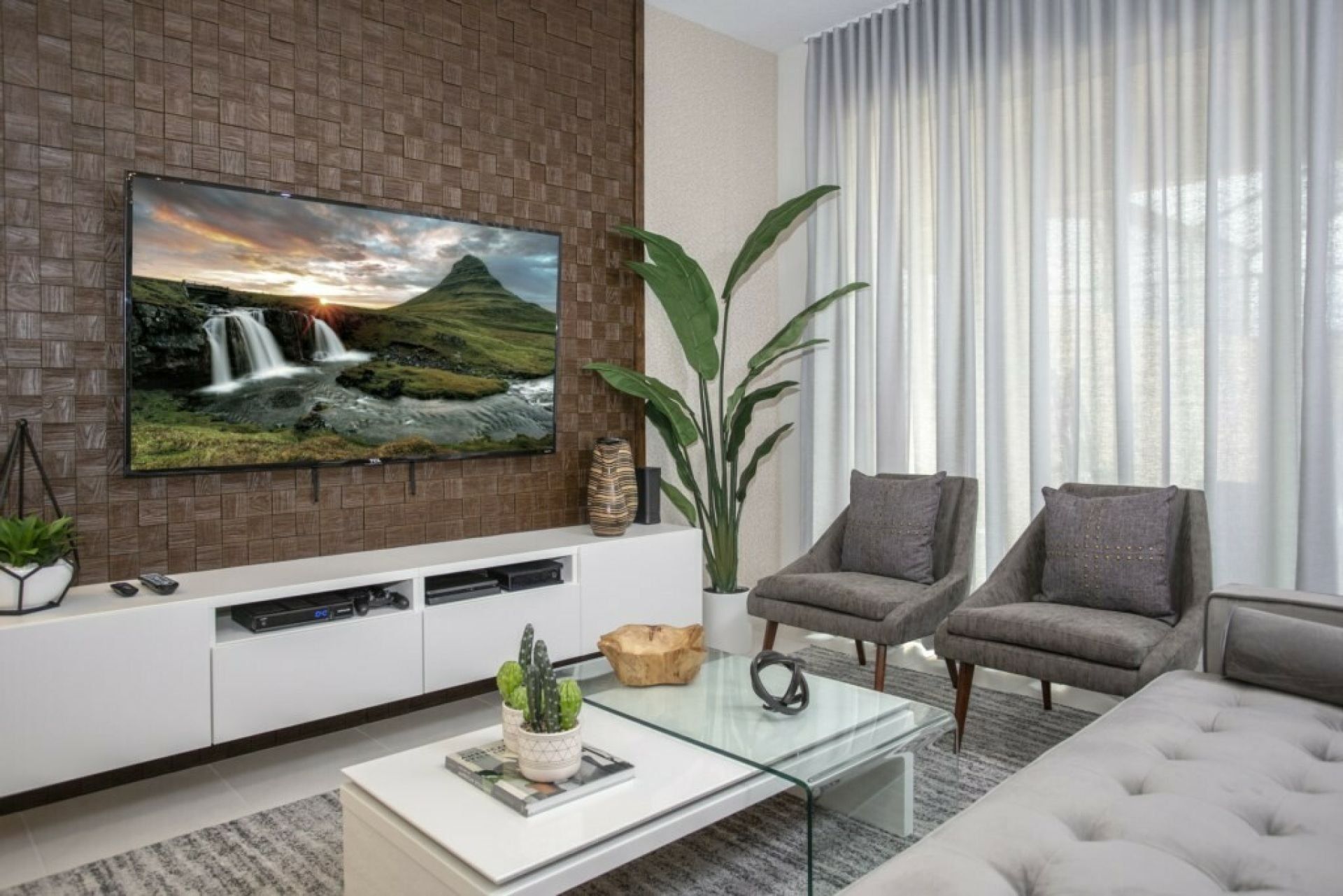 A modern living room with a flat-screen TV mounted on a brick wall, two grey armchairs, a glass coffee table, and a large plant.
