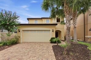 A two-story house with a garage, a paved driveway, and a variety of plants and trees. - 5BR Disney-area home | Pool, theater, themed (Kissimmee)