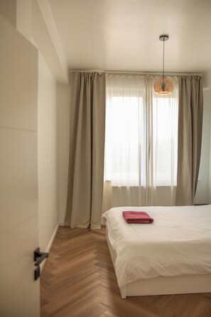 A bedroom with a bed, wooden flooring, a window with curtains, and a hanging light fixture. - Brussels South - Cosy Appartment (Anderlecht (Brussels))