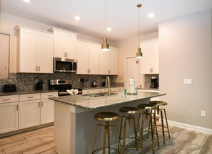 A modern kitchen with white cabinets, a granite countertop, stainless steel appliances, and a central island with bar stools. - Themed 5BR near Disney | Pool, game room (Kissimmee)