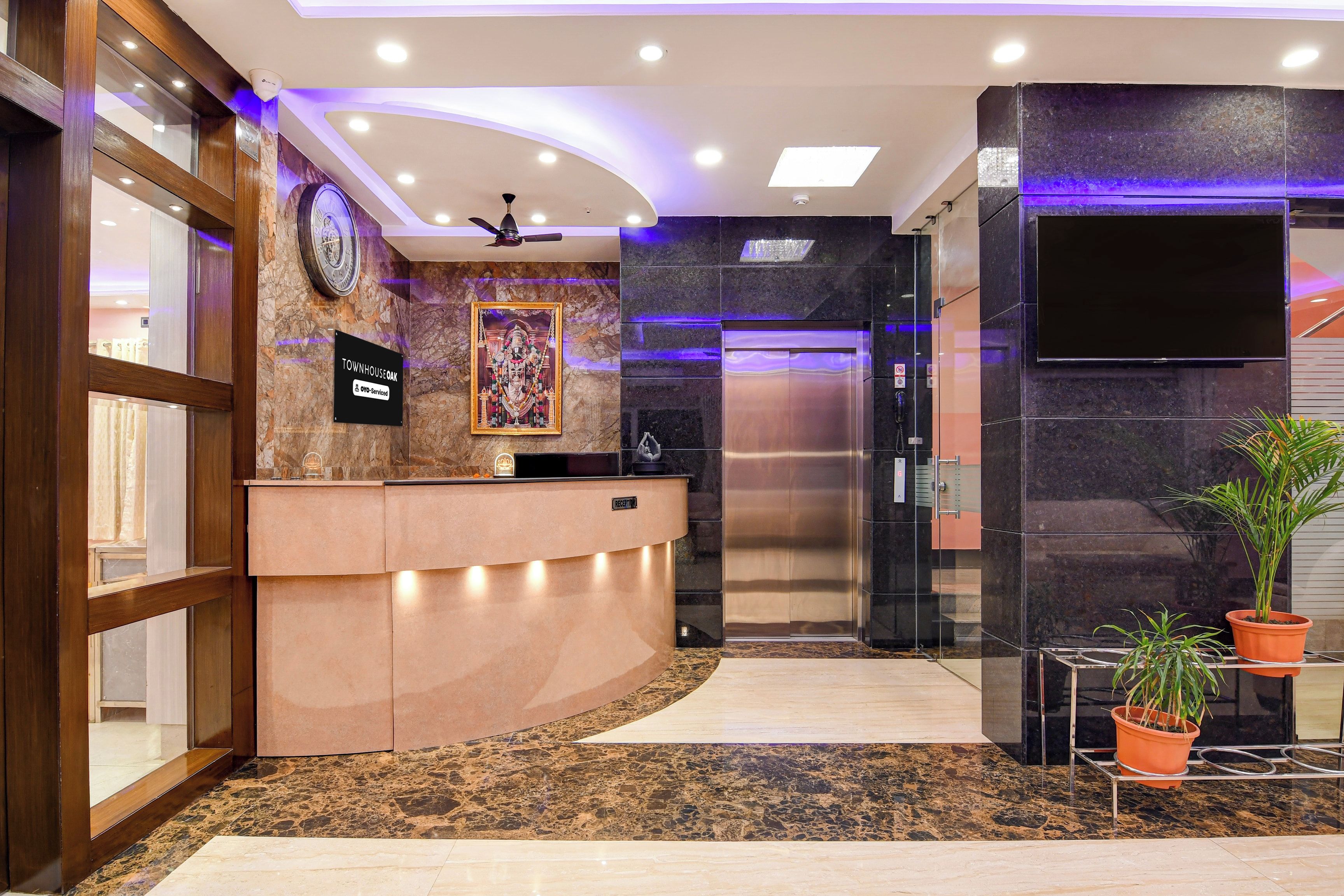 Et hotelværelse med marmorgulv, en receptionsdisk, et ur, et indrammet religiøst billede, en loftsvifte og en moderne elevator.