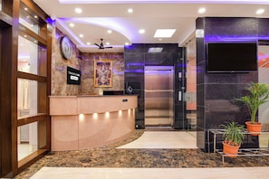 A hotel reception area with a marble floor, a reception desk, a clock, a framed religious picture, a ceiling fan, and a modern elevator.