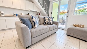 Ein modernes Wohnzimmer mit einem grauen Sofa, passendem Hocker und einer Küchenzeile mit weißen Schränken und einer Kaffeemaschine.