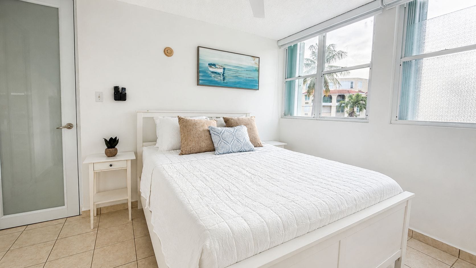 A bedroom with a bed, a nightstand, a wall-mounted light, a painting, and a window with a view of palm trees.