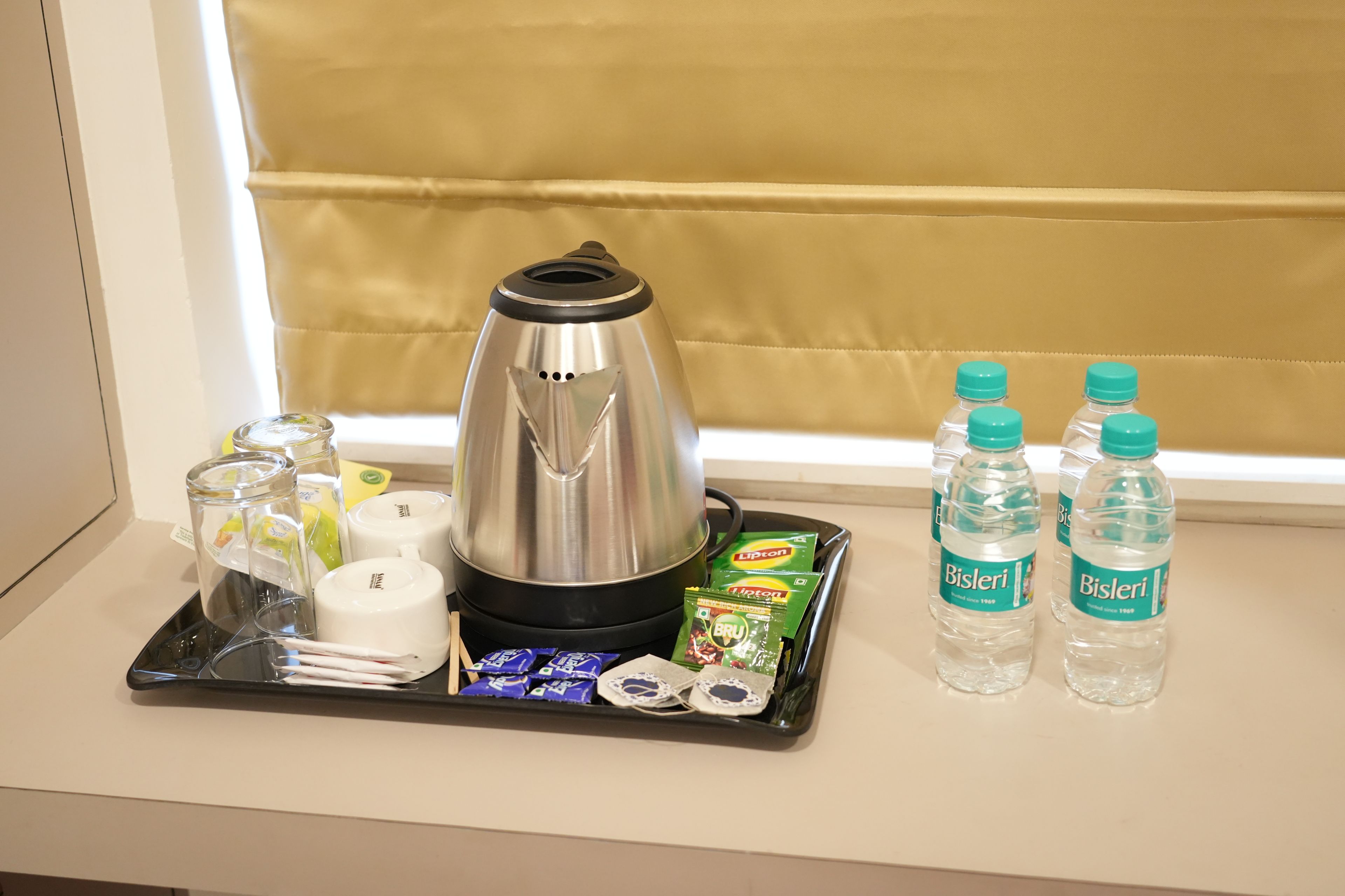 Un plateau contenant une machine à café, des verres, des tasses, des bouteilles d’eau Bisleri et des paquets de collations, posé sur un rebord de fenêtre.