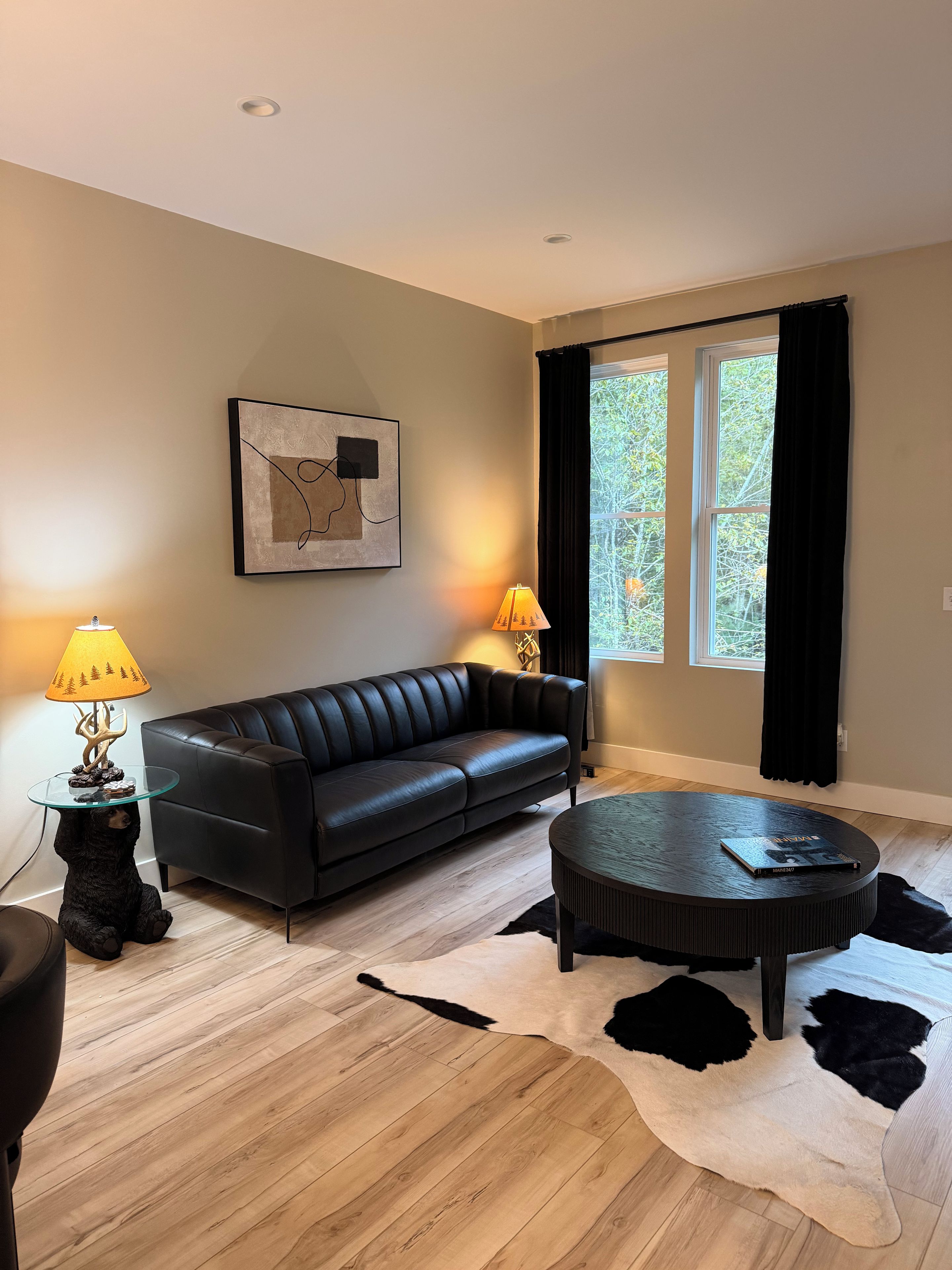 Una sala de estar moderna con un sofá de cuero negro, una mesa de centro de vidrio, una mesita auxiliar redonda, un tapete blanco y negro, y dos lámparas.
