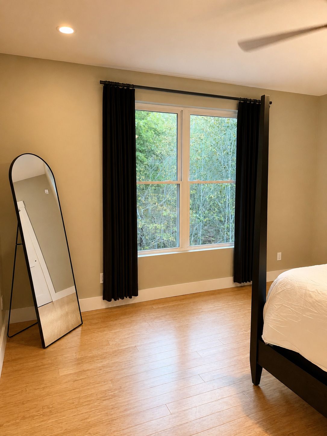 Un dormitorio con cama, ventana con cortinas, espejo de pie y ventilador de techo.