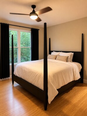 A four-poster bed with white linens, a ceiling fan, and a window with curtains.