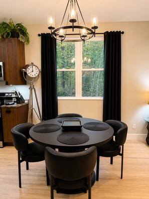 Uma sala de jantar com mesa redonda, cadeiras pretas, lustre, relógio e janela com cortinas.
