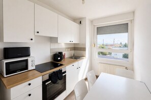 A compact kitchen with white cabinets, a microwave, a coffee maker, and a view of a cityscape through a window. - Bright apartment with terrace and cathedral view (Strasbourg)