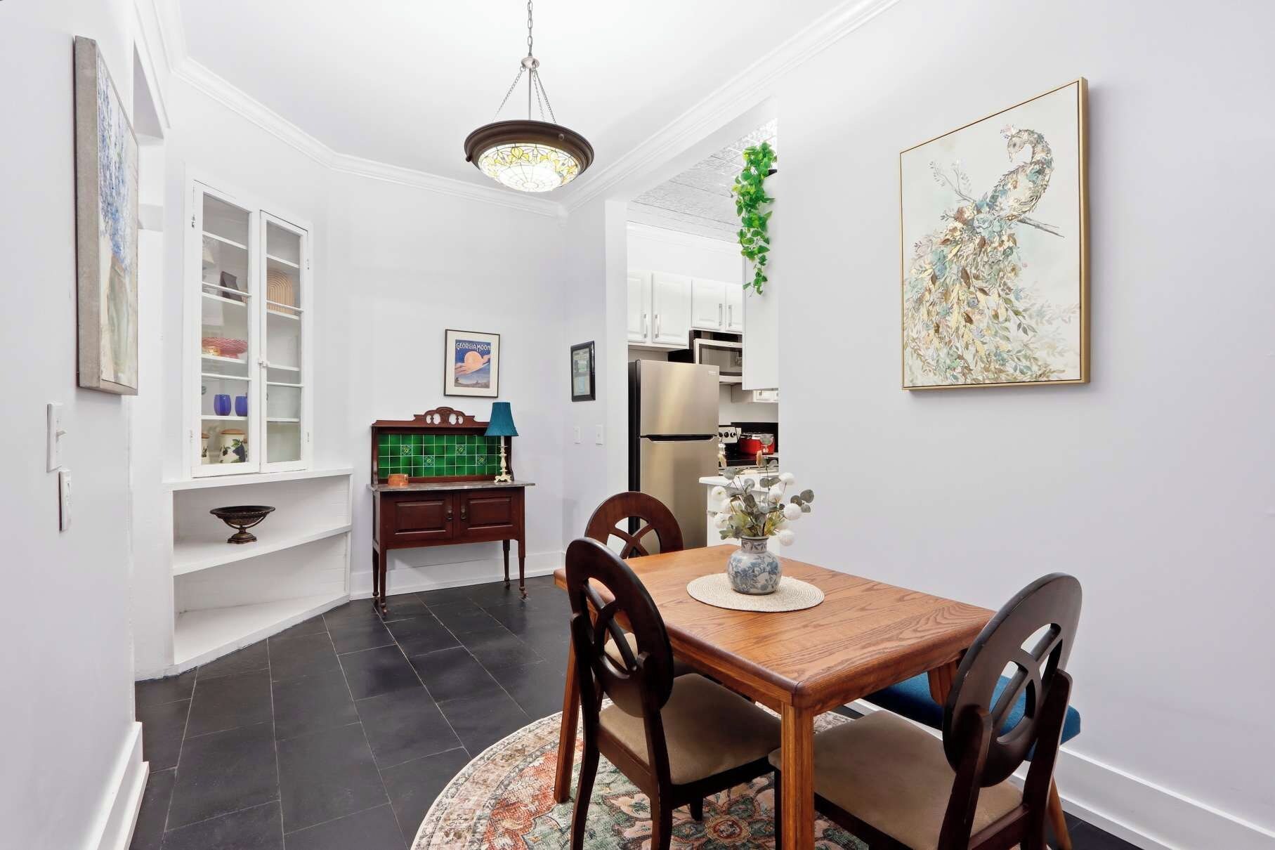 A dining area with a wooden table, chairs, a chandelier, a kitchen in the background, and a wall-mounted artwork.