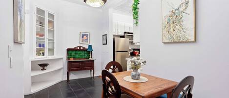 A dining area with a wooden table, chairs, a chandelier, a kitchen in the background, and a wall-mounted artwork.
