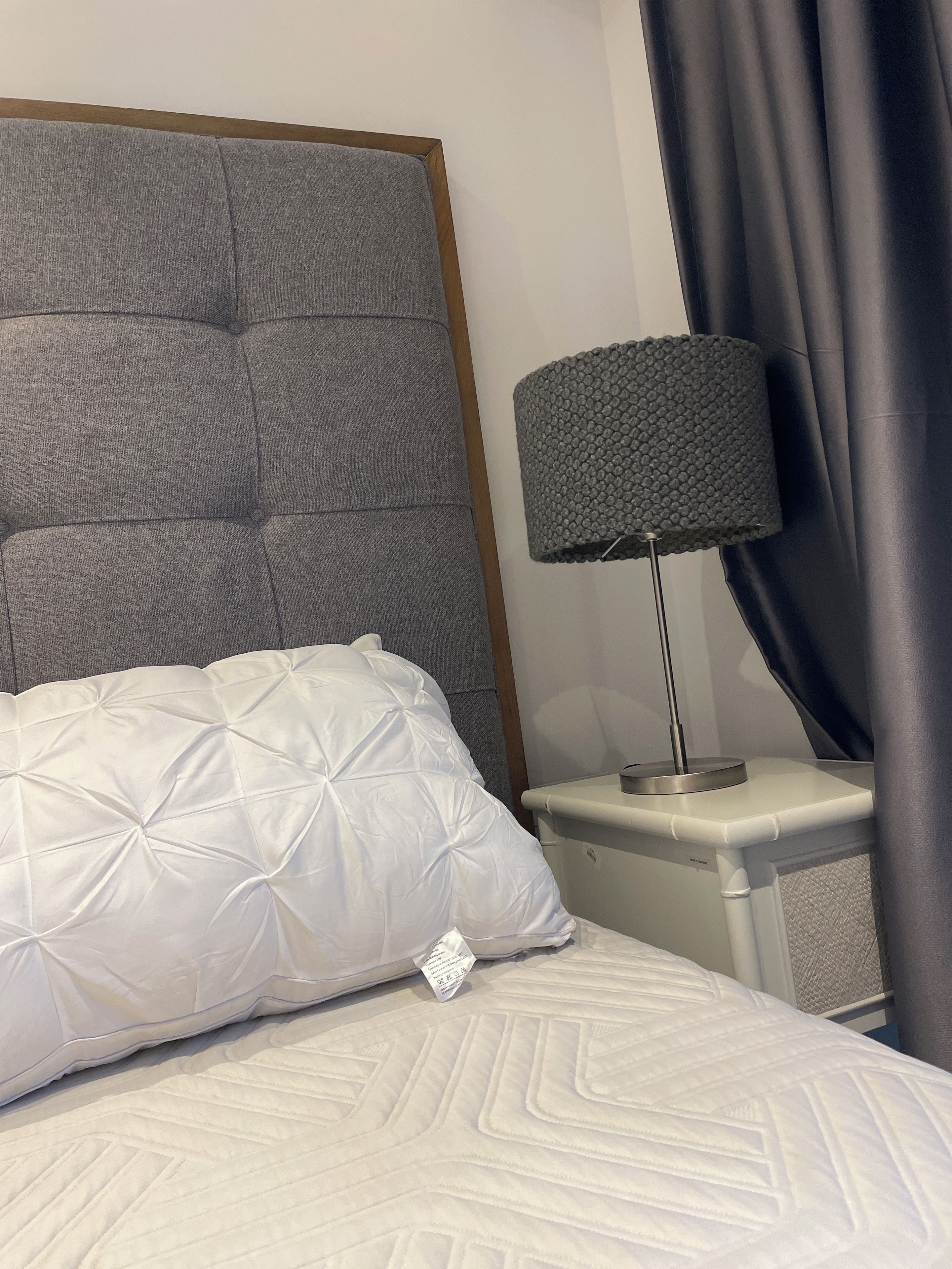 Un lit avec une tête de lit capitonnée, un oreiller blanc, une lampe de chevet et une table de chevet.