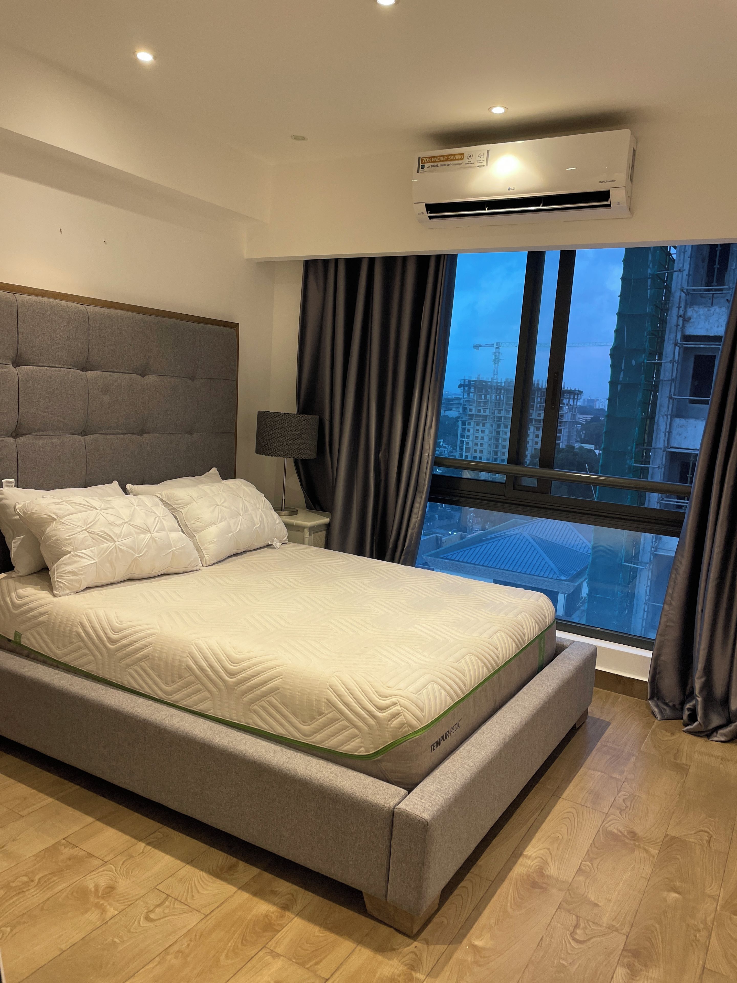 Een moderne slaapkamer met een groot bed, een nachtkastje, een lamp en uitzicht op een stadsbeeld door een raam.