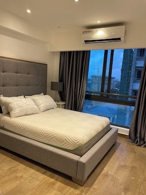 Een moderne slaapkamer met een groot bed, een nachtkastje, een lamp en uitzicht op een stadsbeeld door een raam.