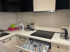 Una cocina con estufa negra, un fregadero, un cajón con utensilios y una encimera con licuadora y hervidor.