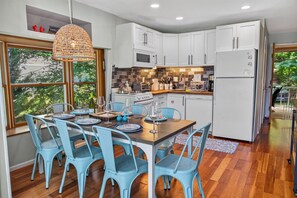 A modern kitchen with white cabinets, a dining table with blue chairs, and a large window overlooking greenery. - Corrotoman River Retreat - Dock, Kayaks, Shuffleboard & Views (Lancaster)