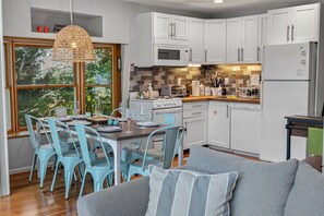 A modern kitchen with white cabinets, a microwave, a stove, and a dining area with blue chairs and a table set for two. - Corrotoman River Retreat - Dock, Kayaks, Shuffleboard & Views (Lancaster)